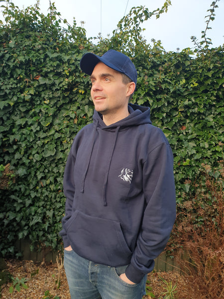 Photo of man wearing a navy trucker's cap with The Cally Collection logo in navy on the front panel.   The man is also wearing a navy hoody with The Cally Collection logo in white in the top left corner.