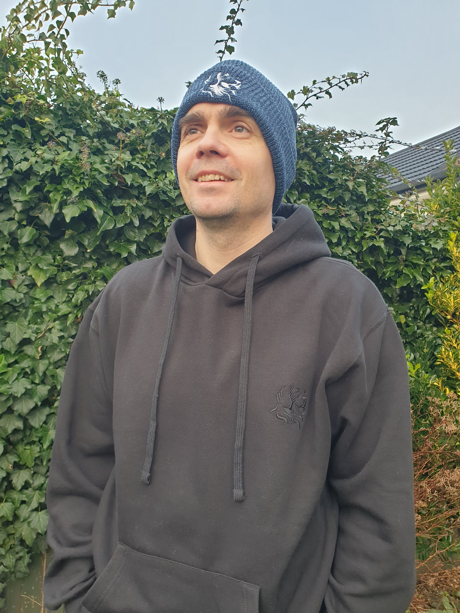 Photo of man wearing a beanie, which is a speckled blue colour, with The Cally Collection logo in white on the front. The man is also wearing a black hoodie with The Cally Collection logo in black in the top left corner.