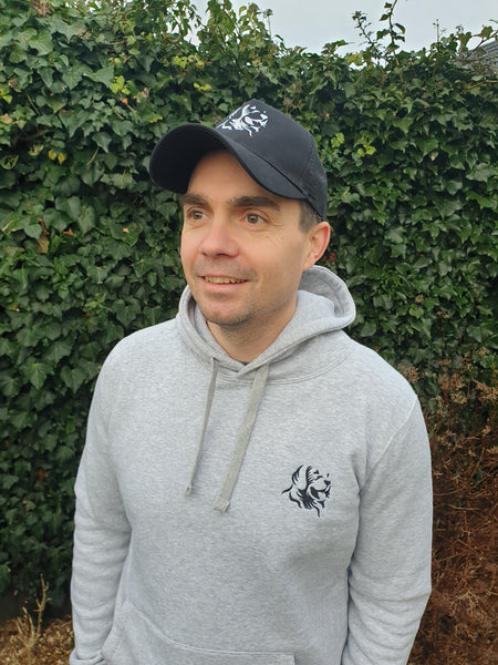 Photo of man wearing a black trucker's cap with The Cally Collection logo in white on the front panel.   The man is also wearing a light grey hoody with The Cally Collection logo in black in the top left corner.