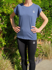 Lady standing with denim coloured t-shirt with cuffed sleeves with The Cally Collection logo in white in the top left hand corner. The lady is also wearing black leggings with the Cally Collection logo in white at the top of the left leg.