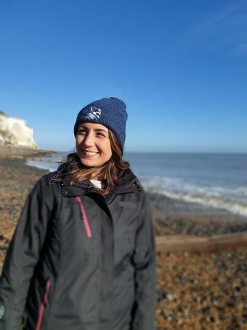 Photo of lady standing on the beach during winter wearing a beanie, which is a speckled blue colour, with The Cally Collection logo in white on the front. 