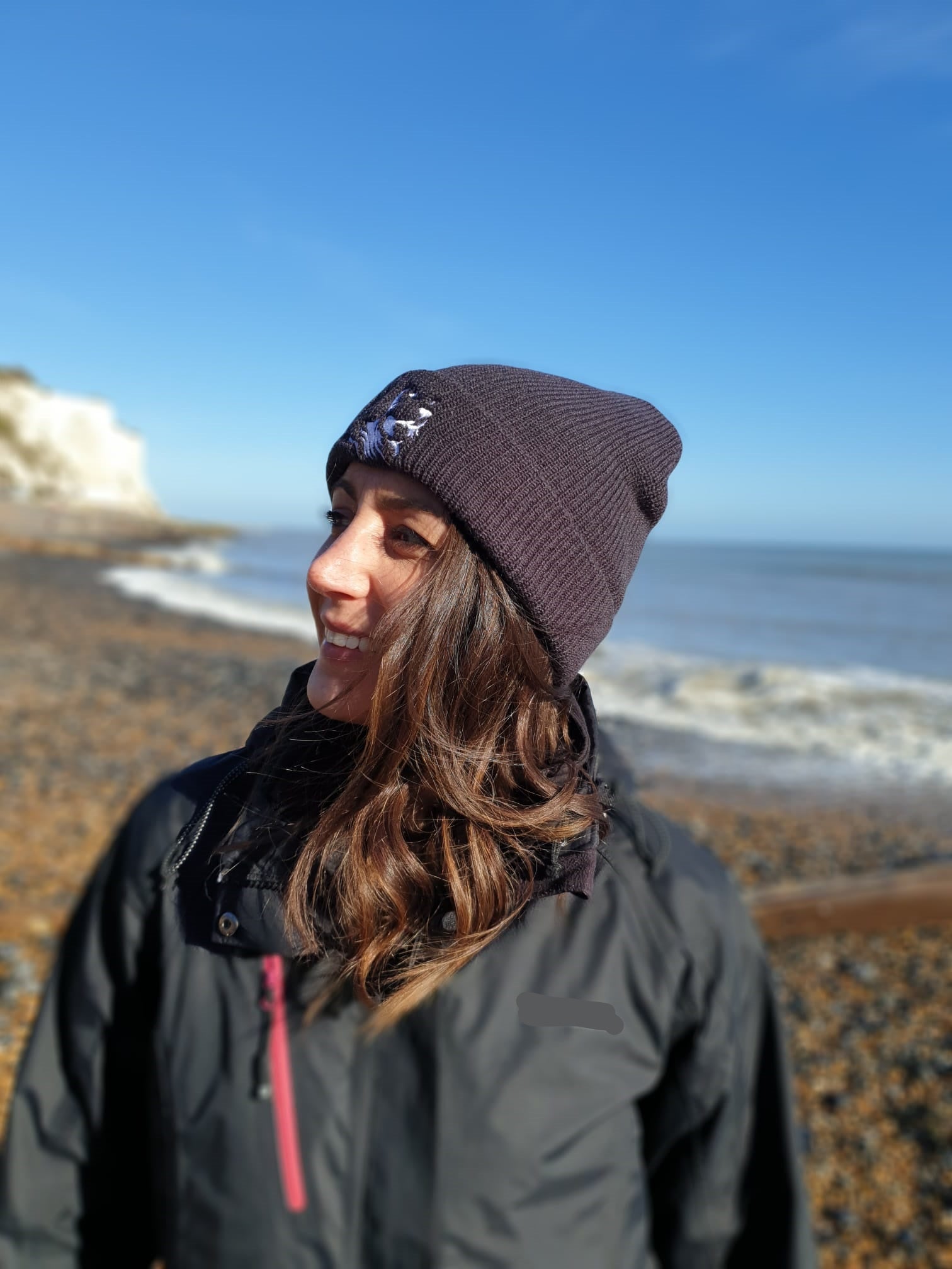 Photo of a lady standing on the beach in winter wearing a dark grey beanie with The Cally Collection logo in white on the front. 