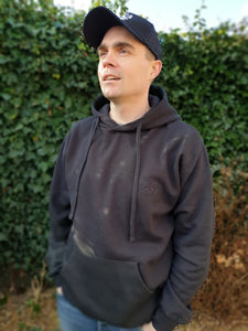 Photo of man wearing a black hoody with The Cally Collection logo in black in the top left corner.    The man is also wearing a black trucker's cap with The Cally Collection logo in white on the front panel.