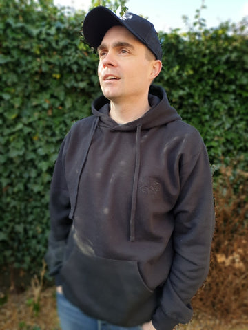 Photo of man wearing a black hoody with The Cally Collection logo in black in the top left corner.    The man is also wearing a black trucker's cap with The Cally Collection logo in white on the front panel.