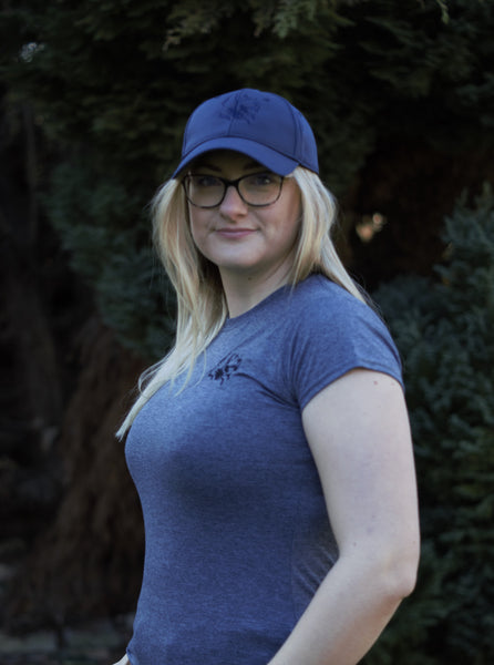 Photo of lady wearing navy air mesh cap with The Cally Collection logo on the front panel.   The lady is also wearing a grey fitted t-shirt with The Cally Collection logo in the top left hand corner.