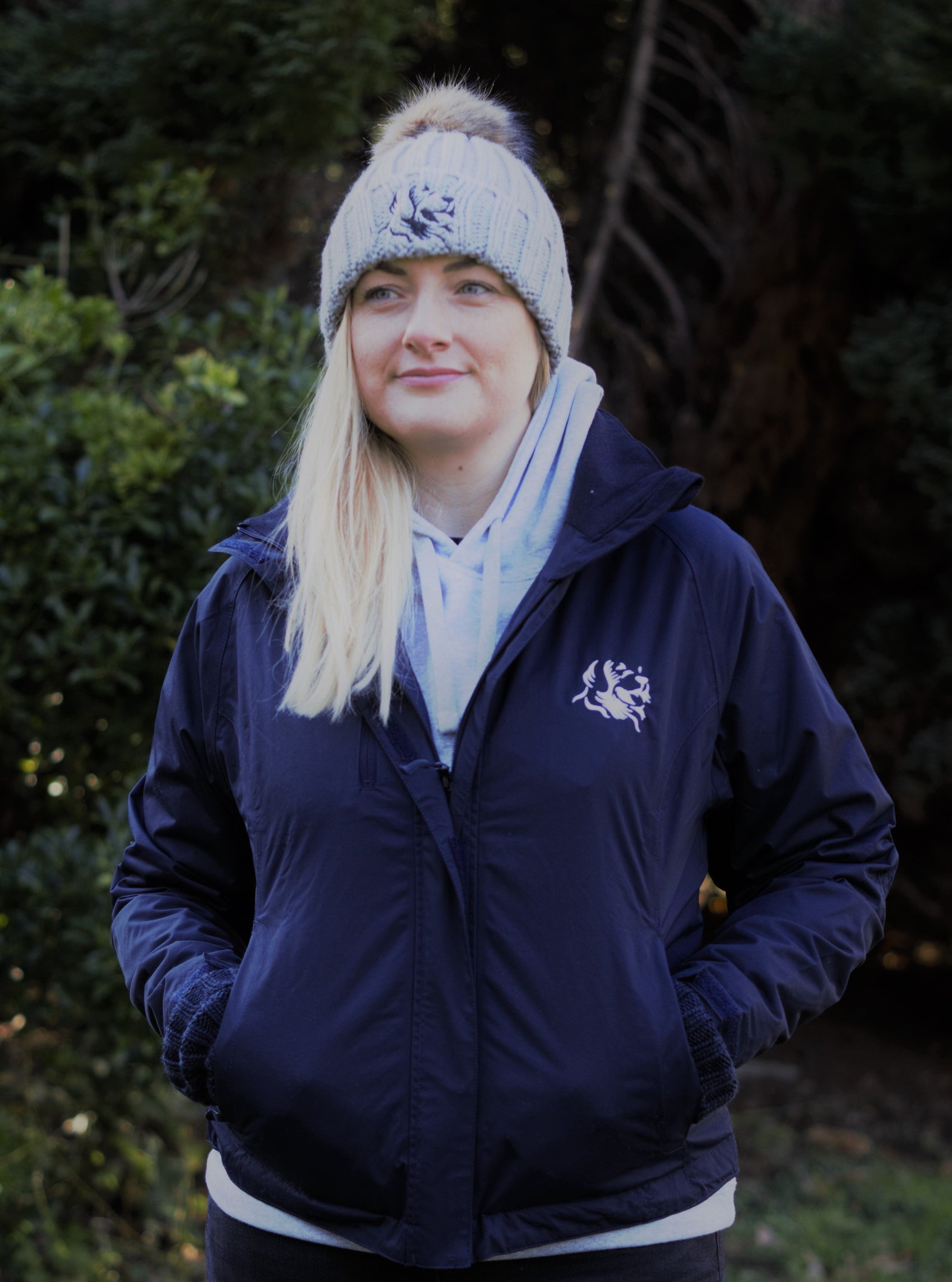 Photo of lady wearing a light grey hoody from The Cally Collection, with a navy waterproof jacket with The Cally Collection logo in white in the top left corner. The lady is also wearing a light grey chunky beanie with a pom pom on the top and The Cally Collection logo in black at the front.