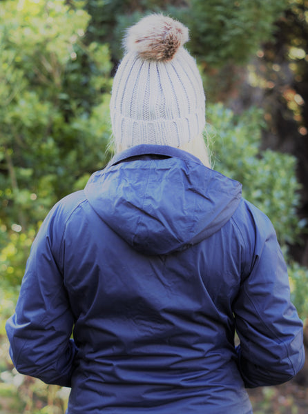 Photo of back of lady wearing a navy waterproof jacket from The Cally Collection . The lady is also wearing a light grey chunky beanie with a pom pom on the top from The Cally Collection.