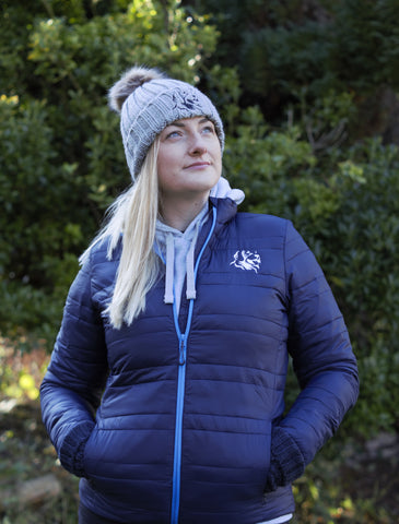 Photo of lady wearing light grey chunky beanie with brown pom pom on the top and The Cally Collection logo at the front in black.   The lady is also wearing a navy soft padded jacket with The Cally Collection logo in white in the top left corner.