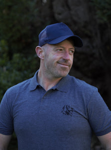 Photo of man wearing a navy trucker's cap with The Cally Collection logo in navy on the front panel.   The man is also wearing a dark grey polo shirt with The Cally Collection logo in black in the top left corner.