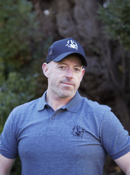 Photo of man wearing a black trucker's cap with The Cally Collection logo in white on the front panel.   The man is also wearing a dark grey polo shirt with The Cally Collection logo in black in the top left corner.