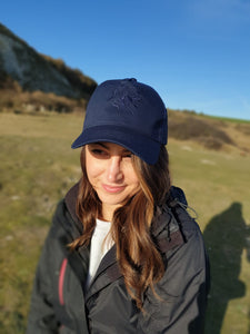 Photo of lady wearing a navy trucker's cap with The Cally Collection logo in navy on the front panel. 